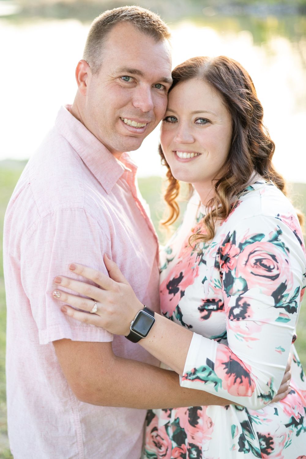 A romantic outdoor portrait showing a couple embracing, with the woman wearing a floral dress and the man in pink shirt.