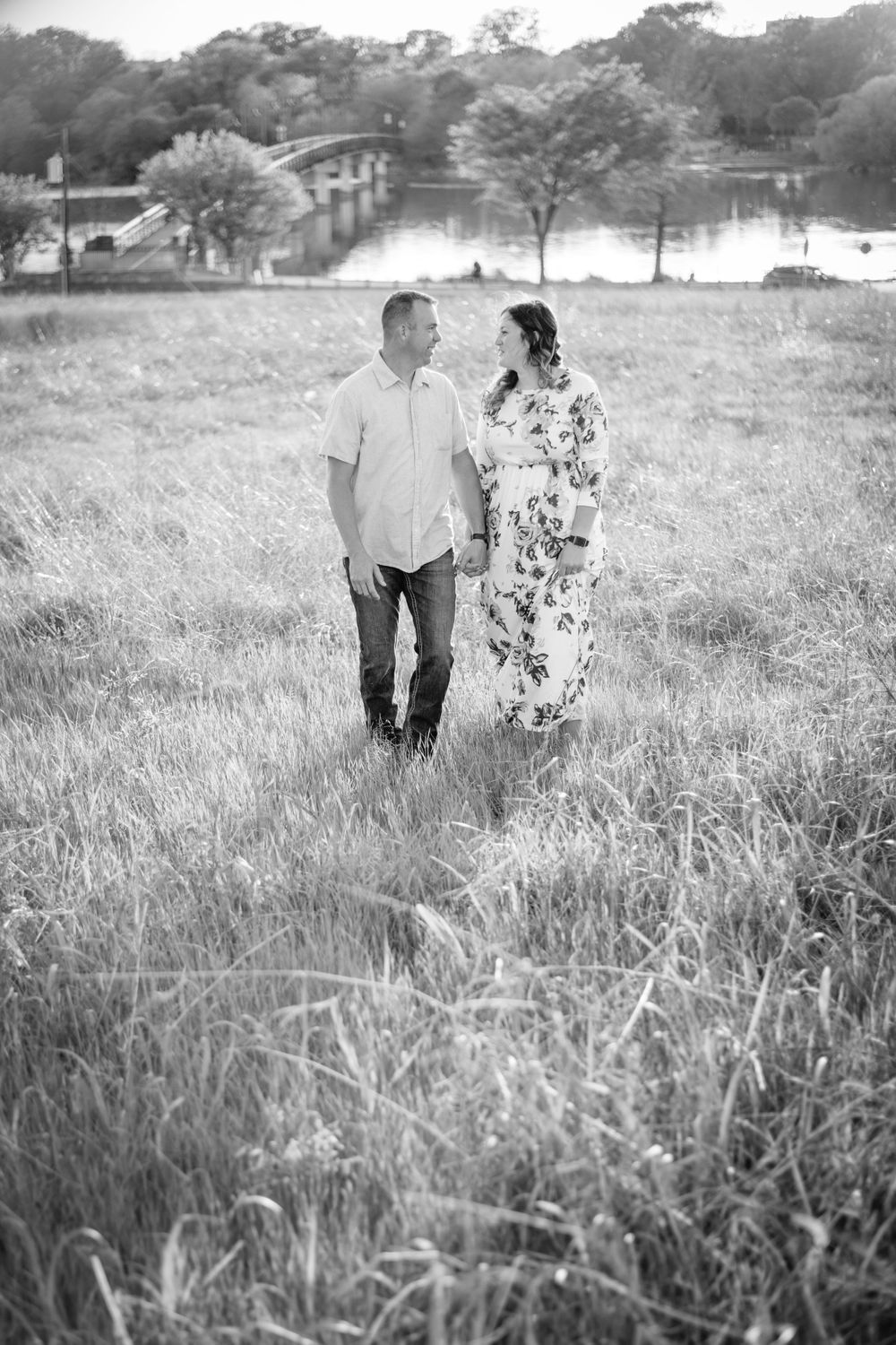 A romantic couple walking through tall grass at sunset near a lake.