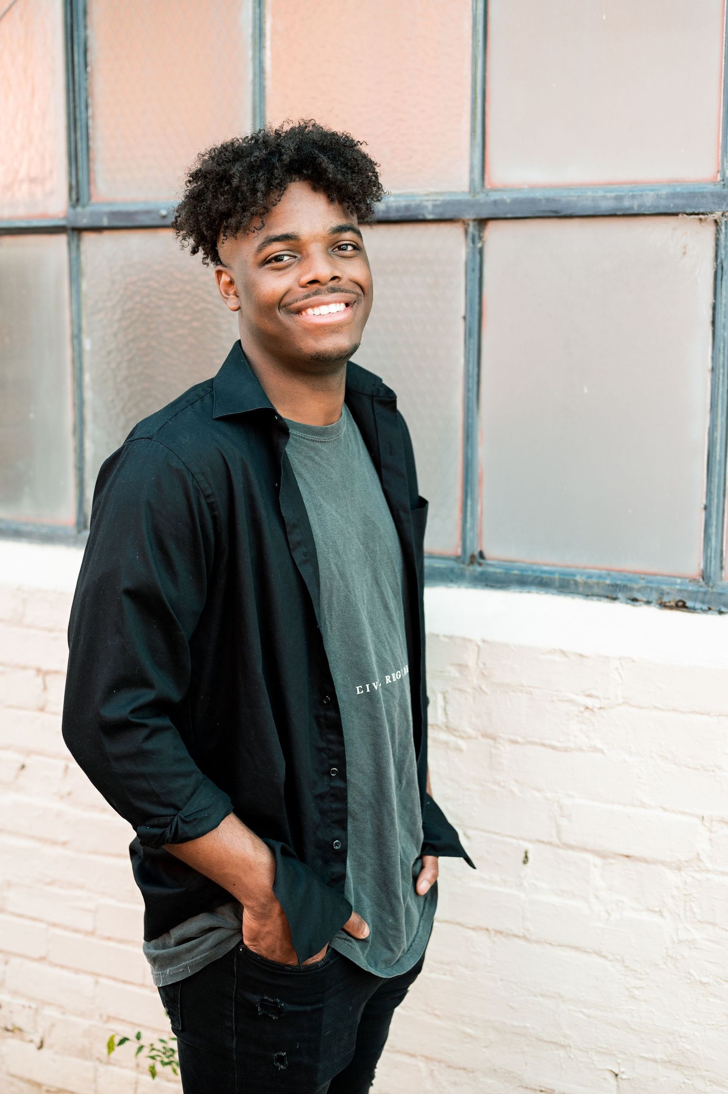 A person in a black jacket and gray shirt poses against a white painted wall during a portrait session.