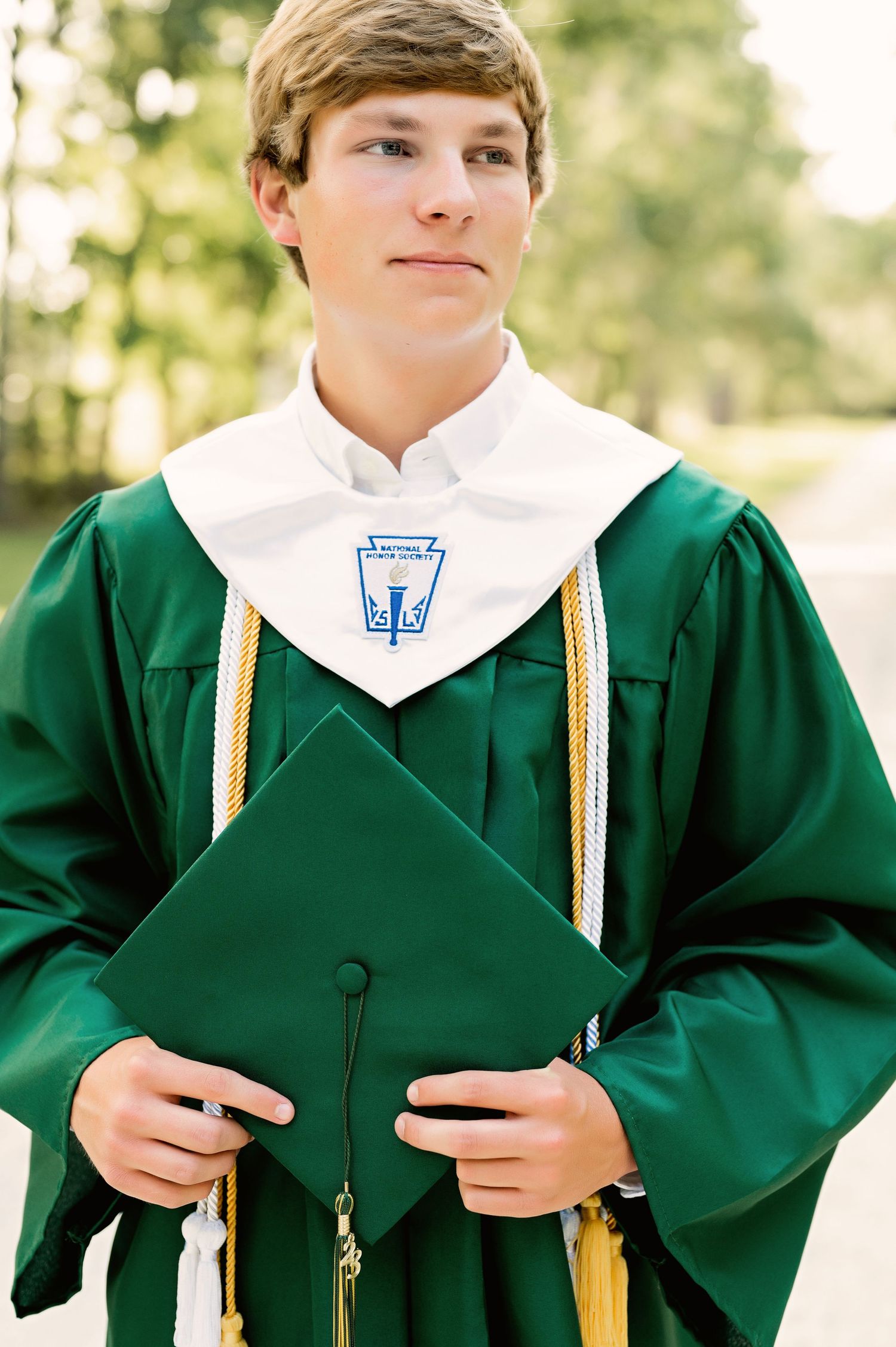 Graduate wearing emerald green academic gown with white collar and gold honor cords in outdoor setting.