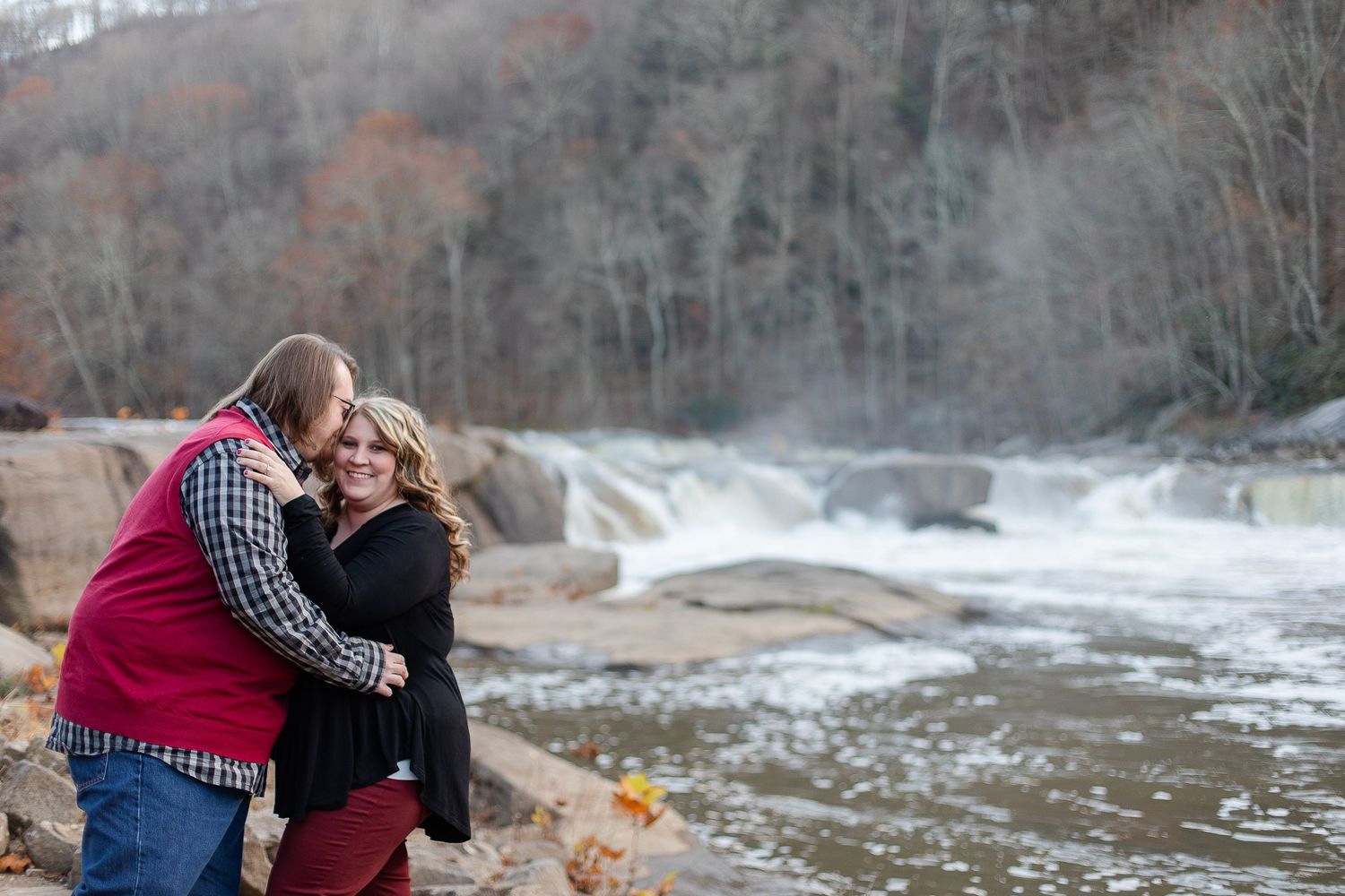 Two people embrace near a rushing waterfall on a cloudy autumn day.