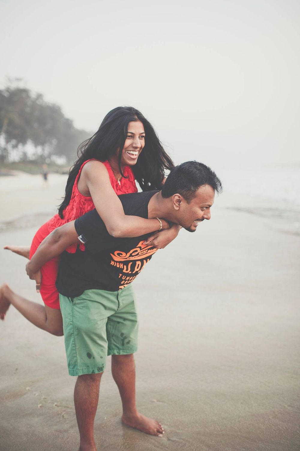 A couple plays and laughs together on a foggy beach, with one person giving the other a piggyback ride in Goa, India