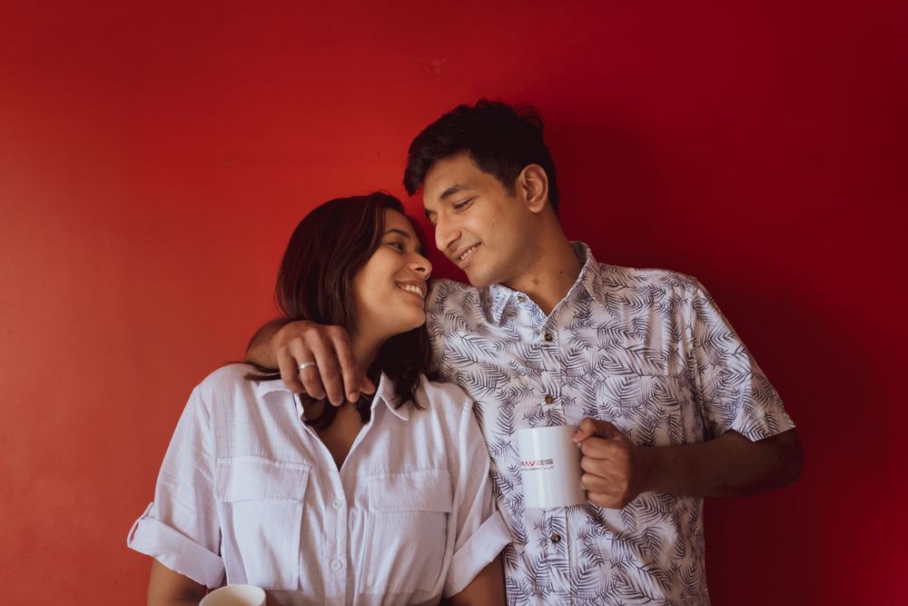 Couple in white shirts sharing an intimate moment against a red backdrop while one holds a coffee mug.