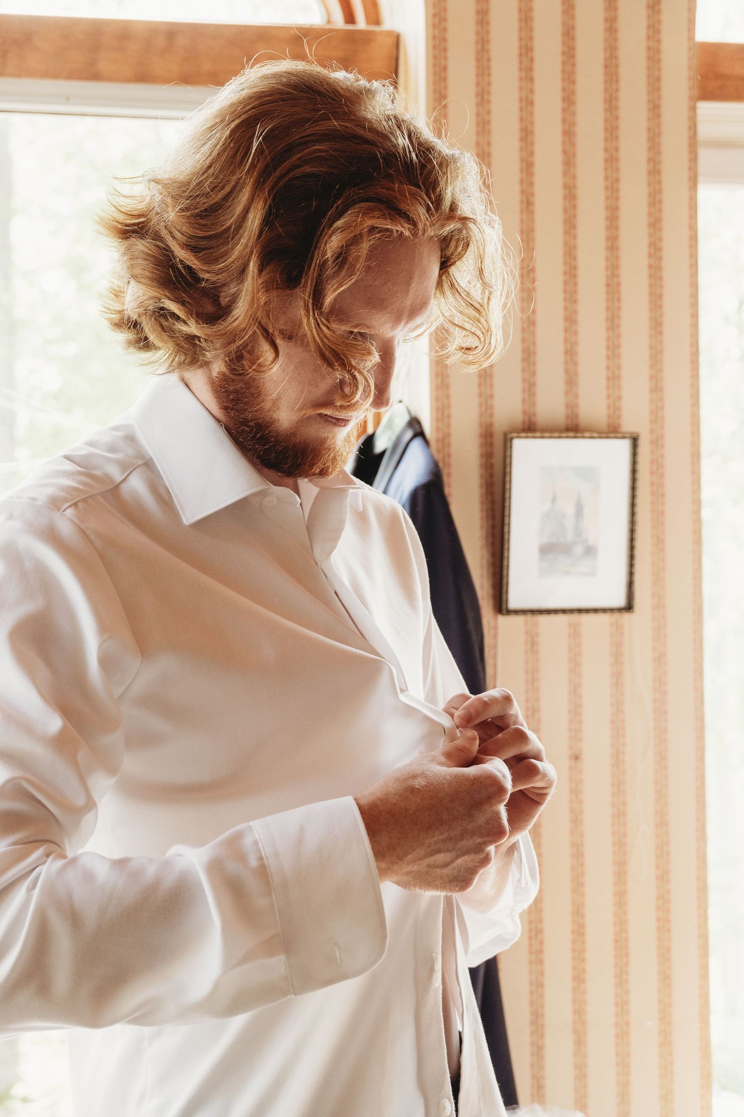 A person wearing a white dress shirt buttoning up their shirt standing by a window with striped wallpaper.