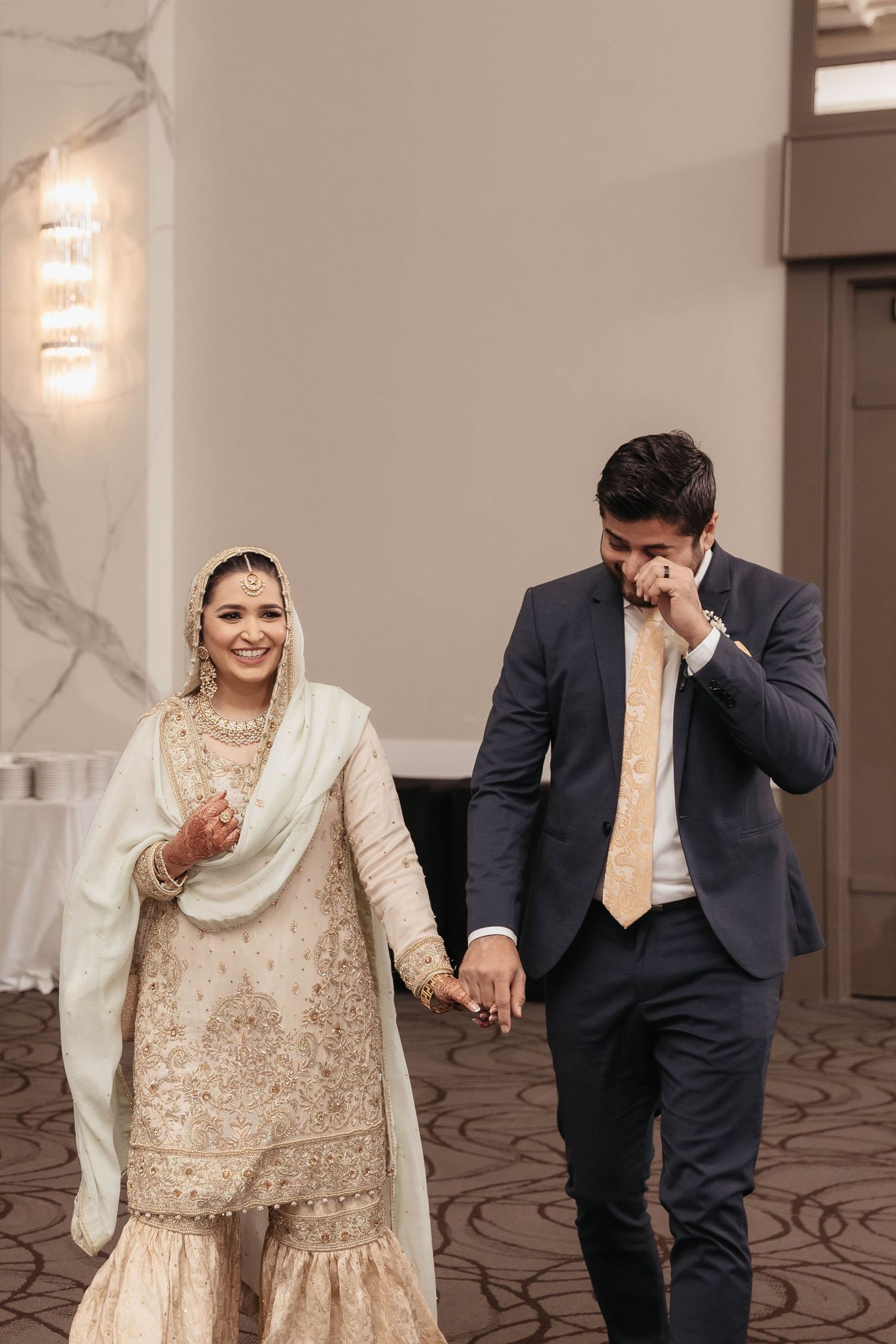 Couple in traditional South Asian wedding attire walking hand in hand indoors.