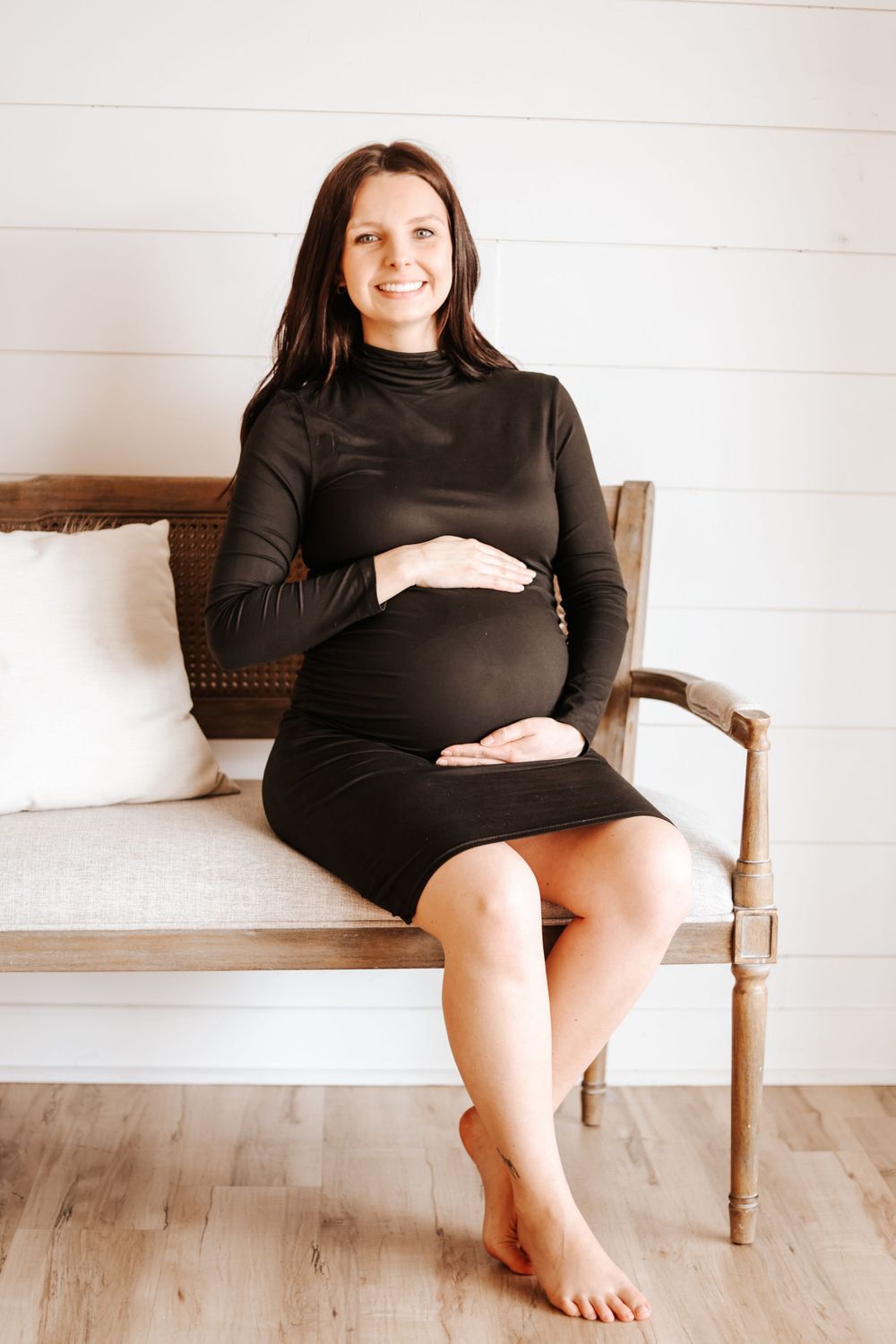 Pregnant woman in black dress relaxes on wooden chair against white wall.