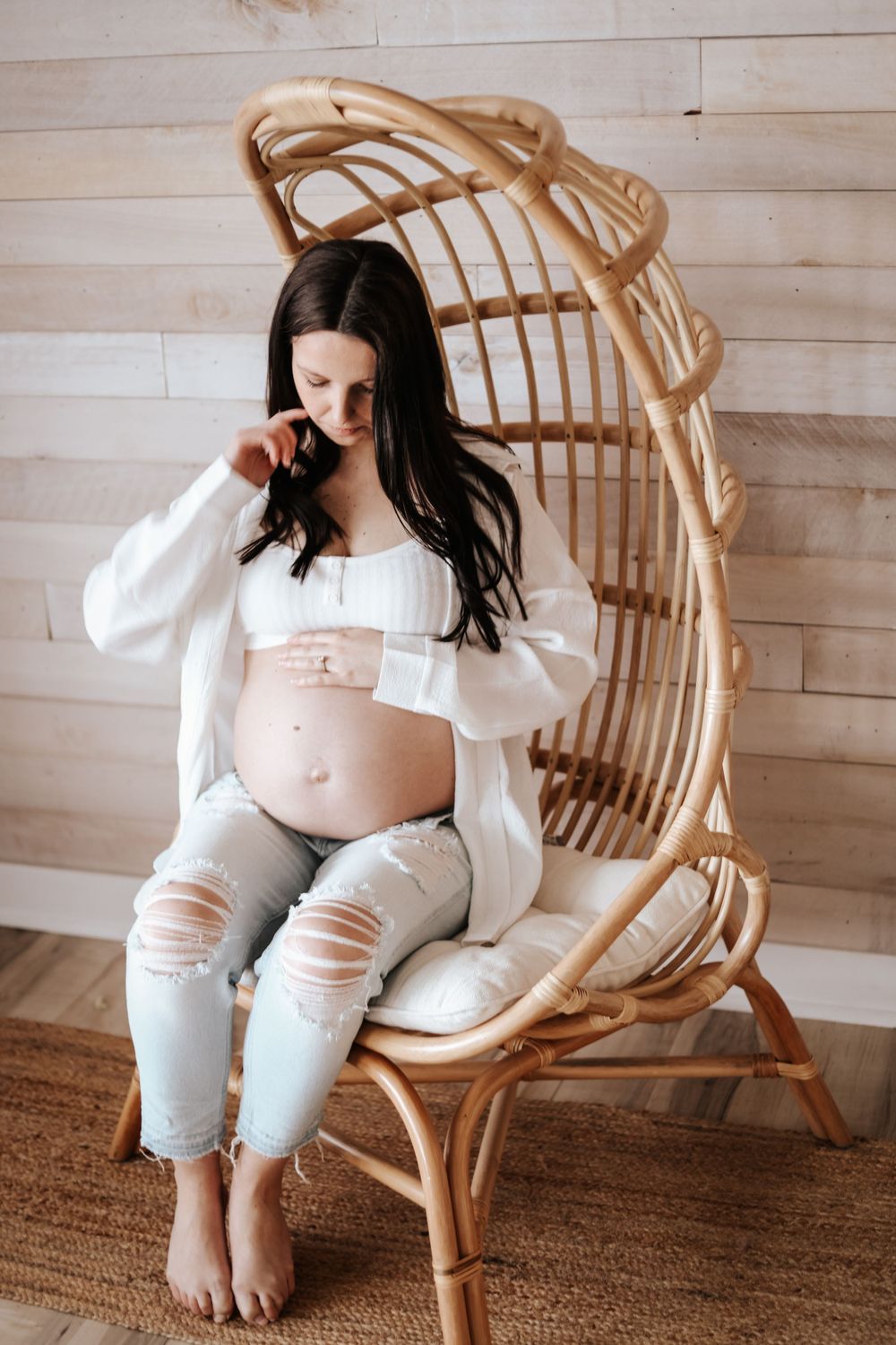 Expectant mother in white shirt and distressed jeans sits in rattan peacock chair against white shiplap wall.