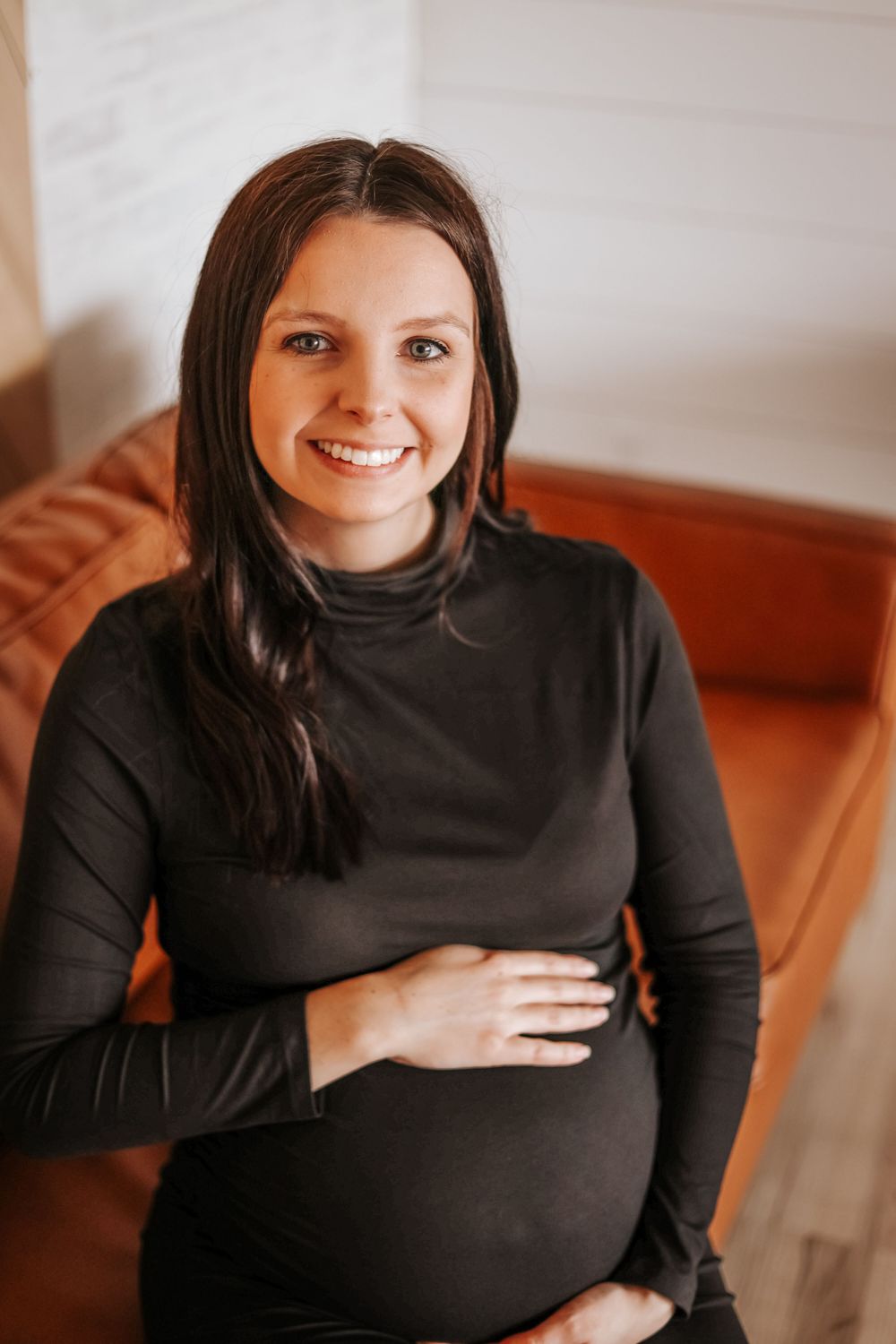 Maternity portrait of an expectant mother in black turtleneck sitting on brown leather chair.