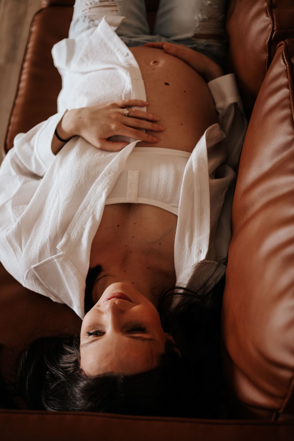 Artistic maternity photo of woman in white shirt reclining on brown leather couch.
