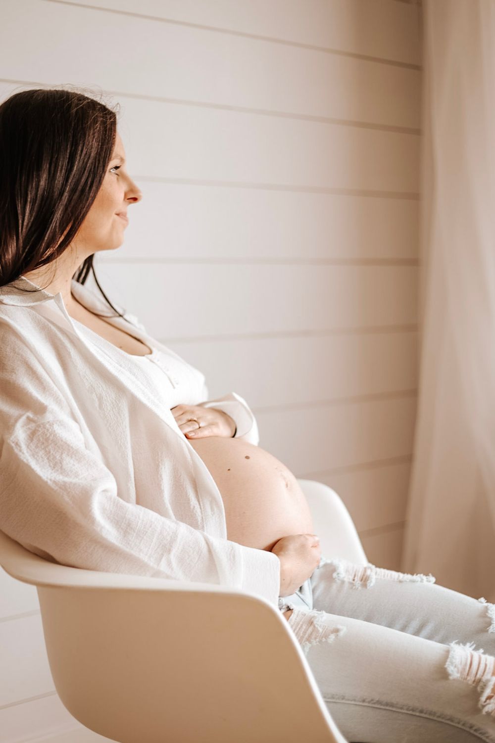 Side profile of pregnant woman in white sitting in modern chair against shiplap wall.