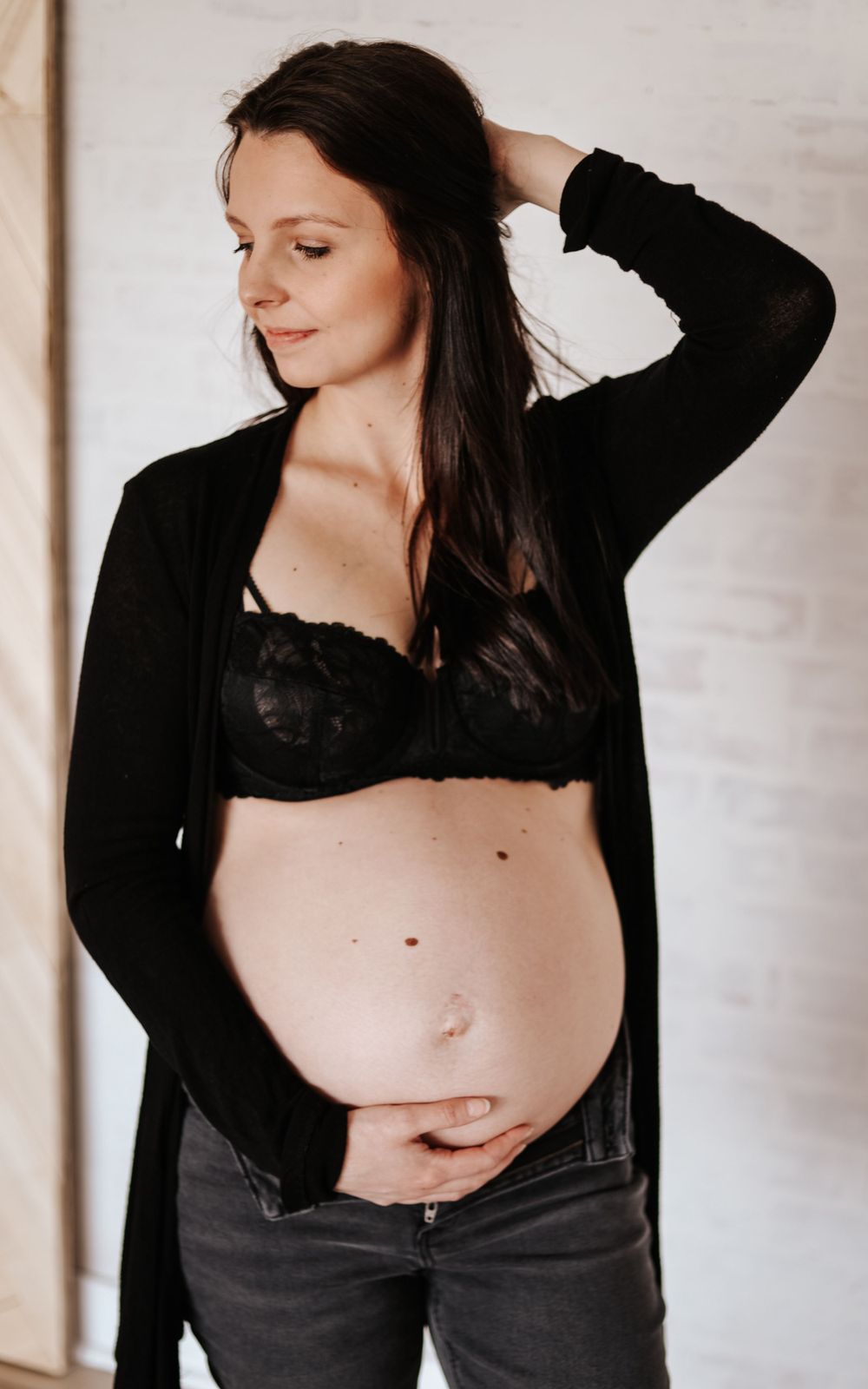 Maternity portrait featuring a pregnant woman in black cardigan and lingerie against white wall.