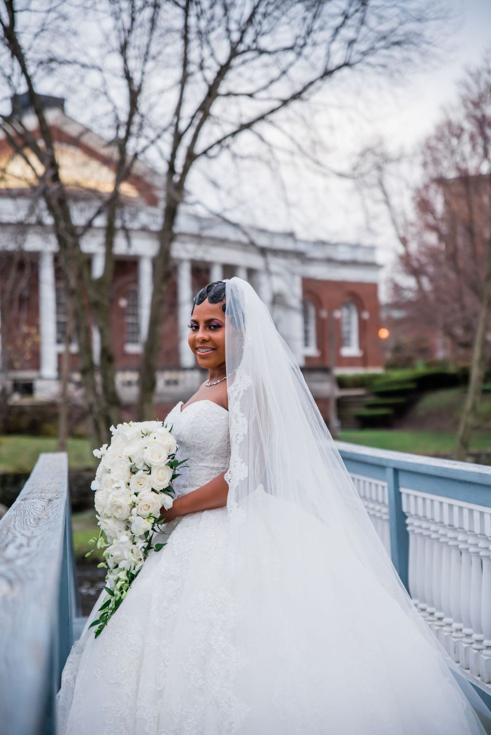 Elegant bride wearing a flowing white ball gown and veil poses on a blue-railed balcony with cascading bouquet.