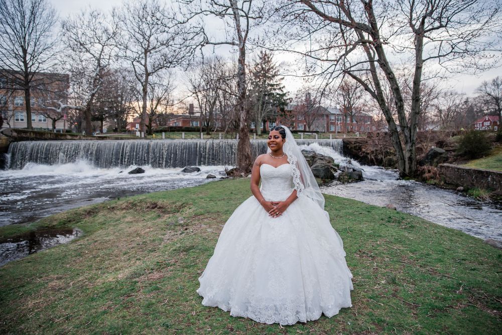 Bride in white ball gown stands by scenic waterfall and landscaped grounds during sunset.