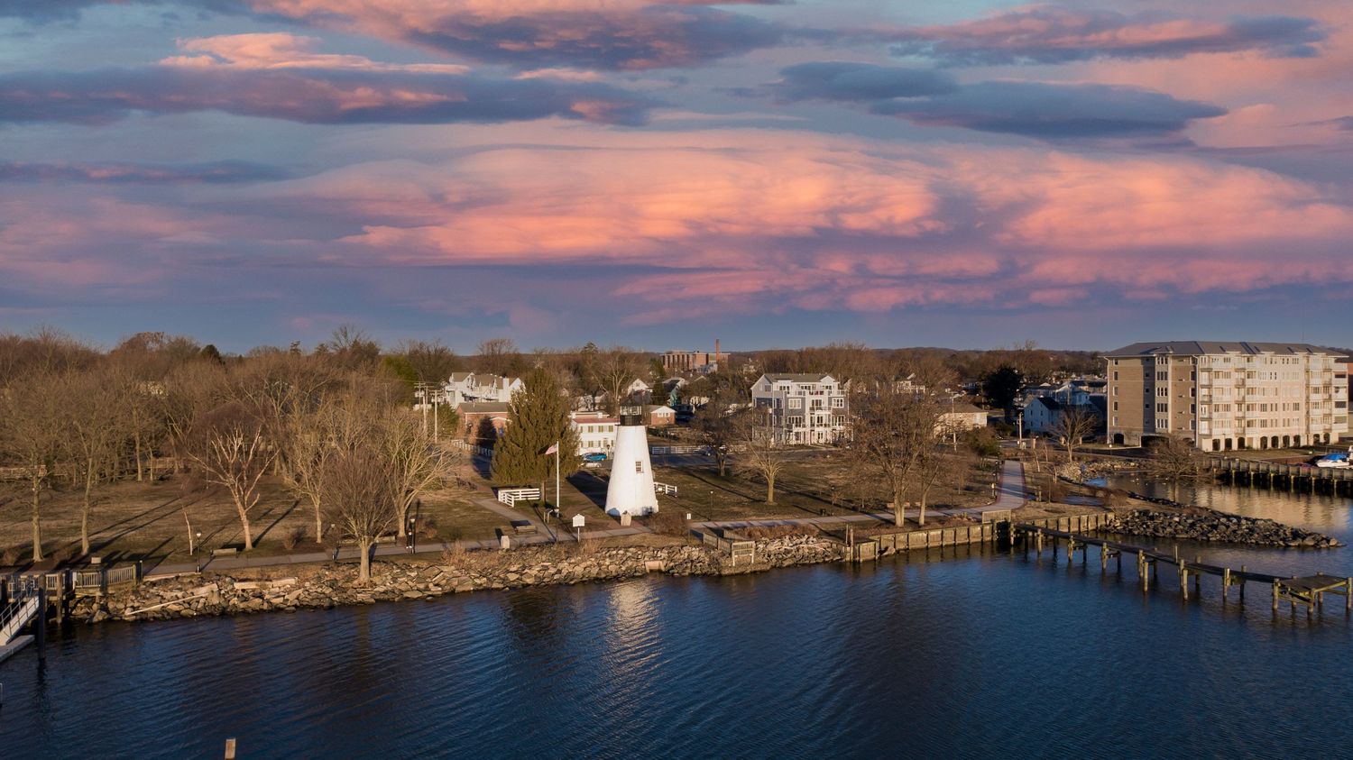 Aerial sunset view of a small coastal town harbor with a white lighthouse and colorful pink clouds.