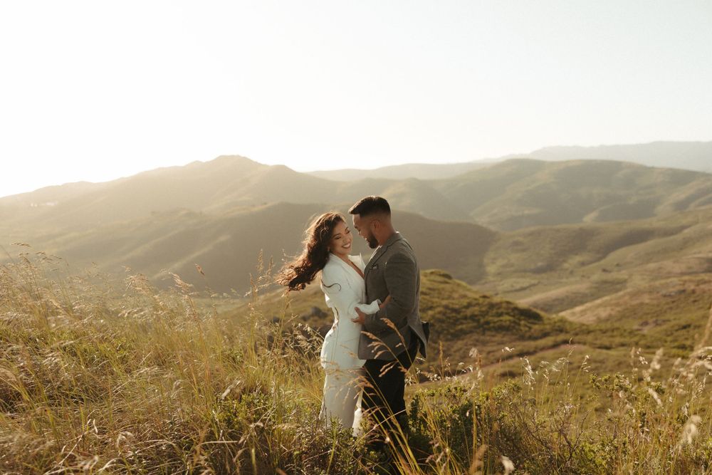 A romantic couple embraces during golden hour on a hilltop overlooking rolling mountains in the distance.