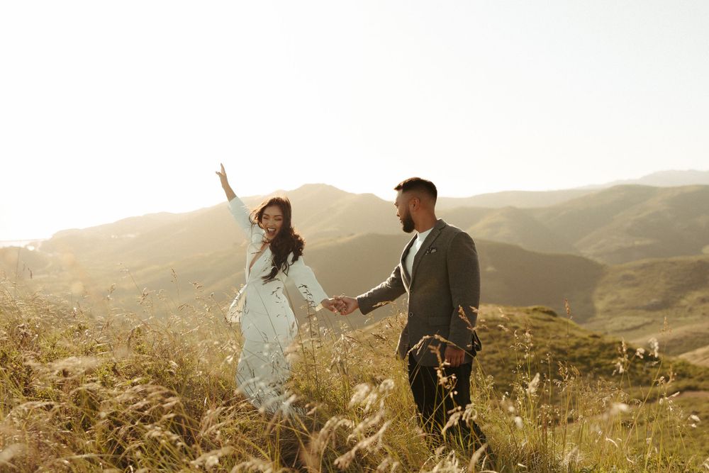 Two people dance joyfully in a grassy field at sunset with mountains in the background.