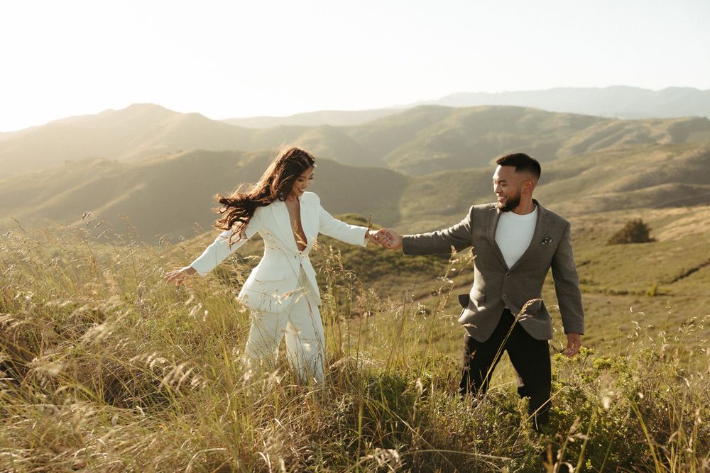 A couple holds hands while spinning playfully in a mountain meadow at sunset.