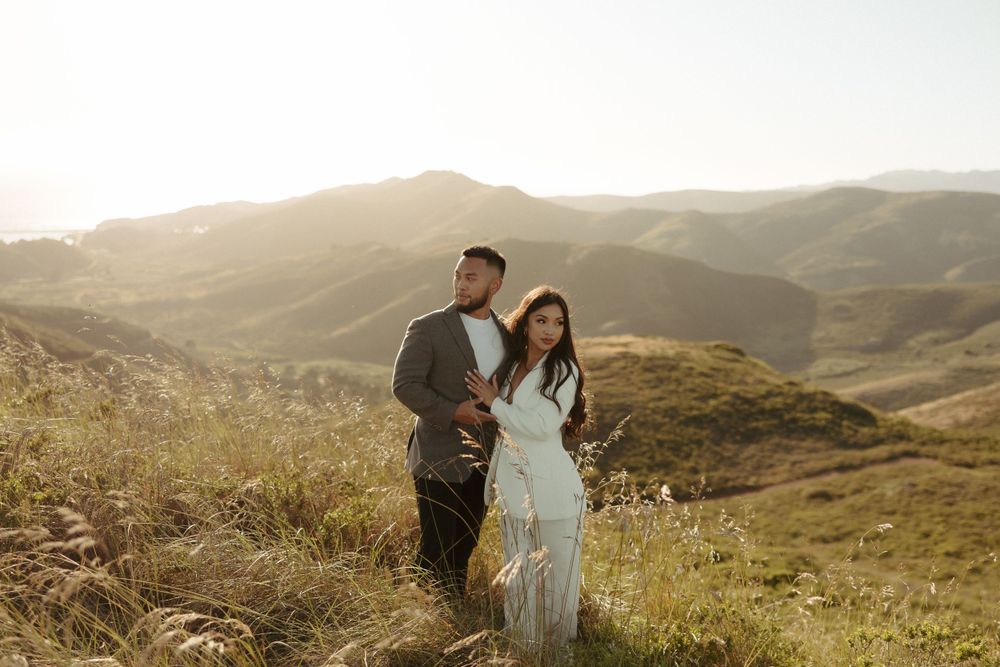 Couple standing together on mountainside trail surrounded by wild grass at golden hour.