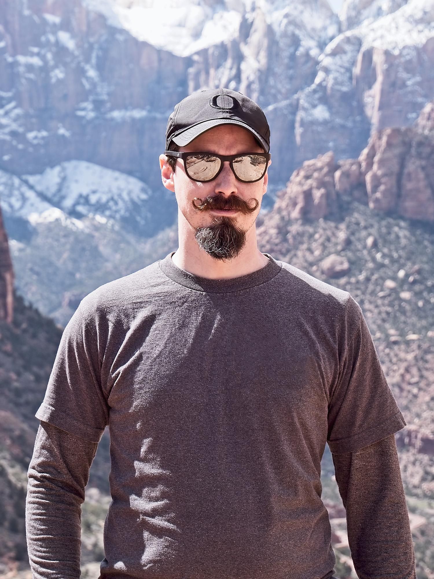 a man in a grey shirt with sunglasses and a curly mustache and goatee in front of a picturesque background