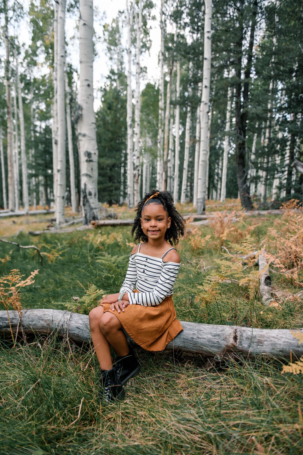 Child in striped shirt poses on fallen log in aspen forest with autumn ferns.