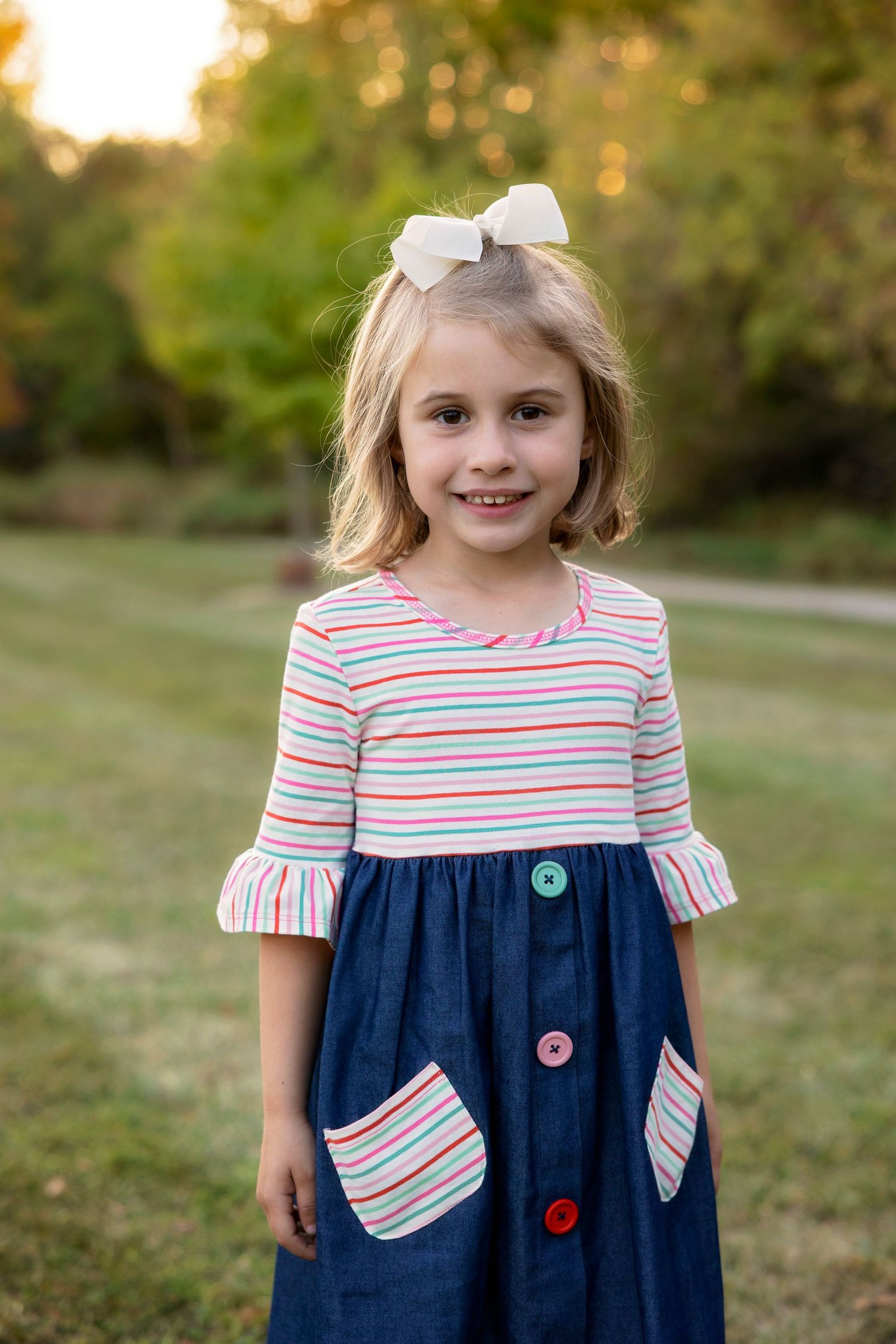 Young child in striped dress with blue skirt and white bow poses outdoors during sunset.