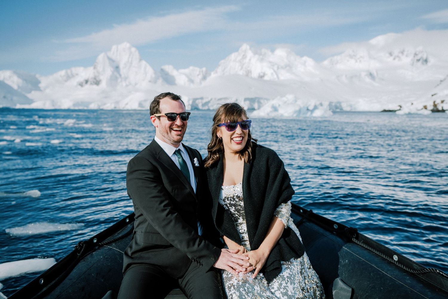 A couple in formal attire sitting in a small boat on blue Antarctic waters with glaciers behind them.