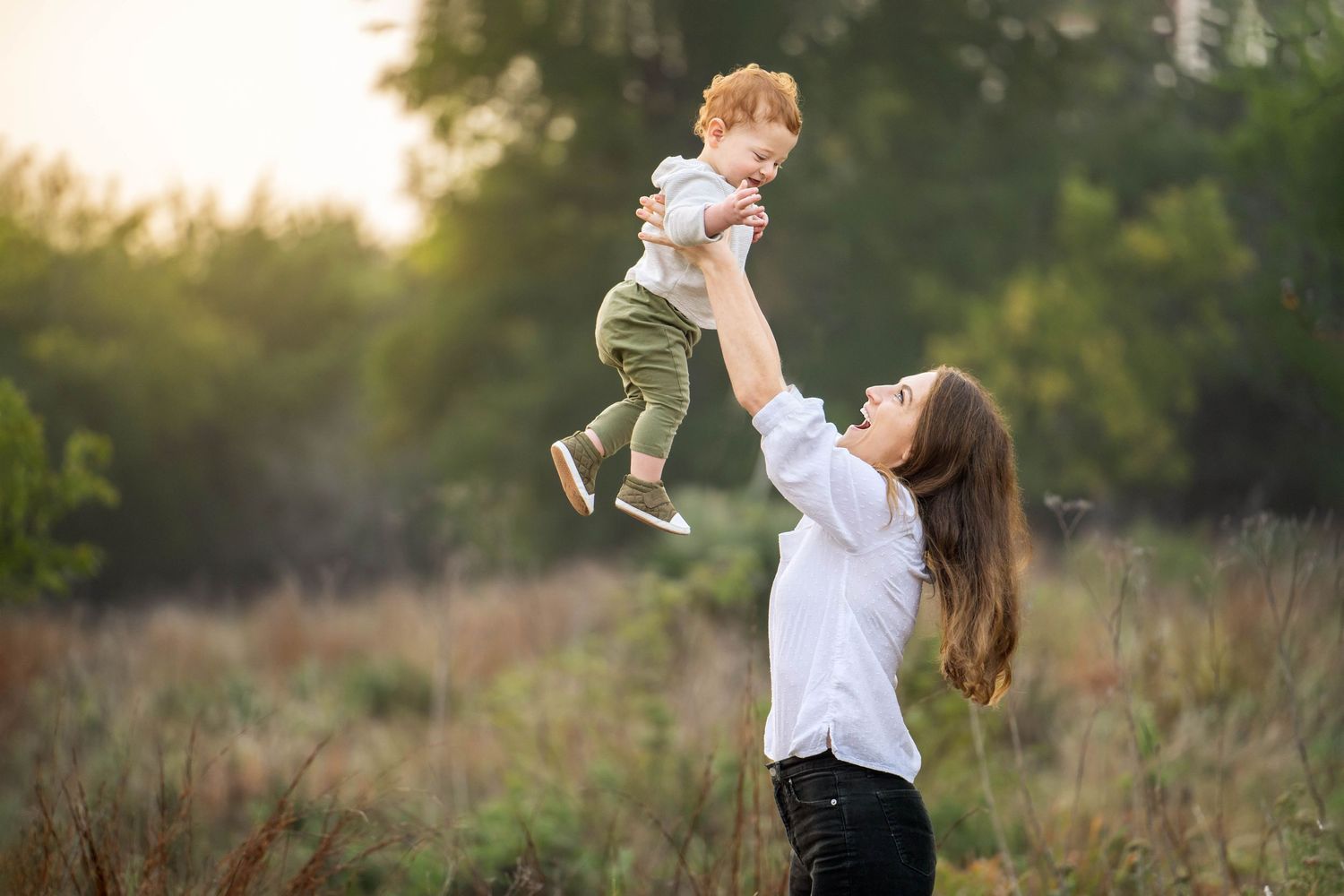 mom in white shirt lifting toddler son with red hair into the air