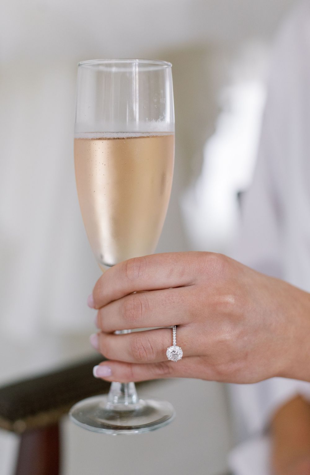 Hand holding champagne flute with sparkling rose wine and engagement ring on finger.