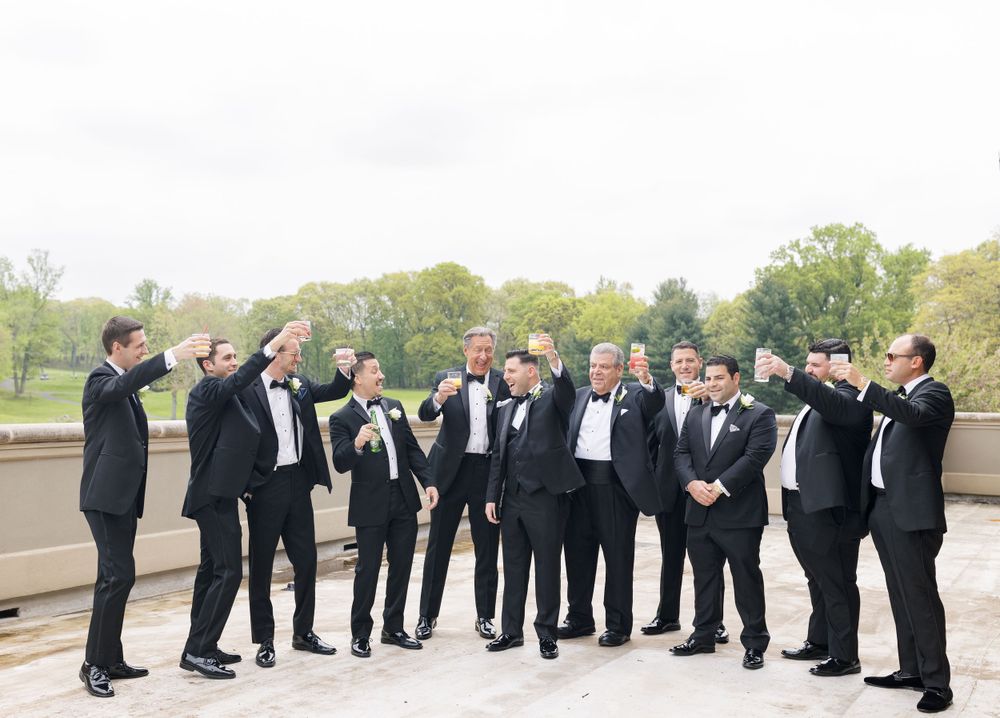 Groomsmen in classic black tuxedos gather outdoors for formal wedding photo.