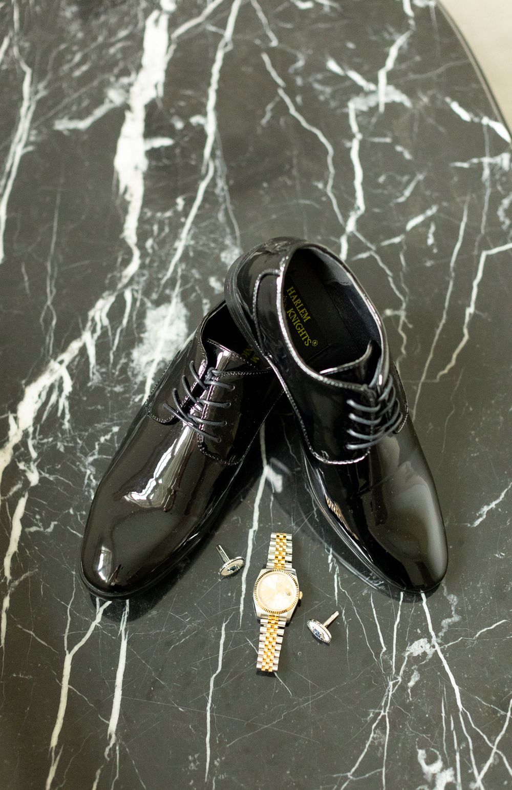 Shiny black dress shoes and gold watch displayed on black marble surface.