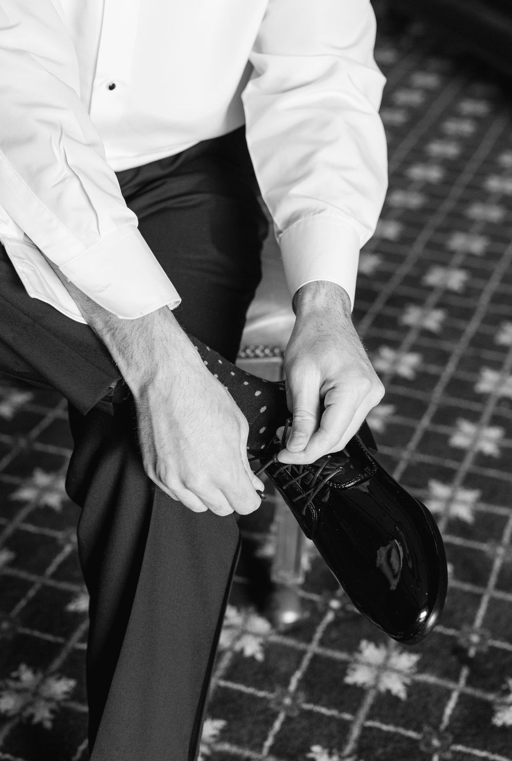 Black and white photo of hands tying formal dress shoe laces on patterned carpet.