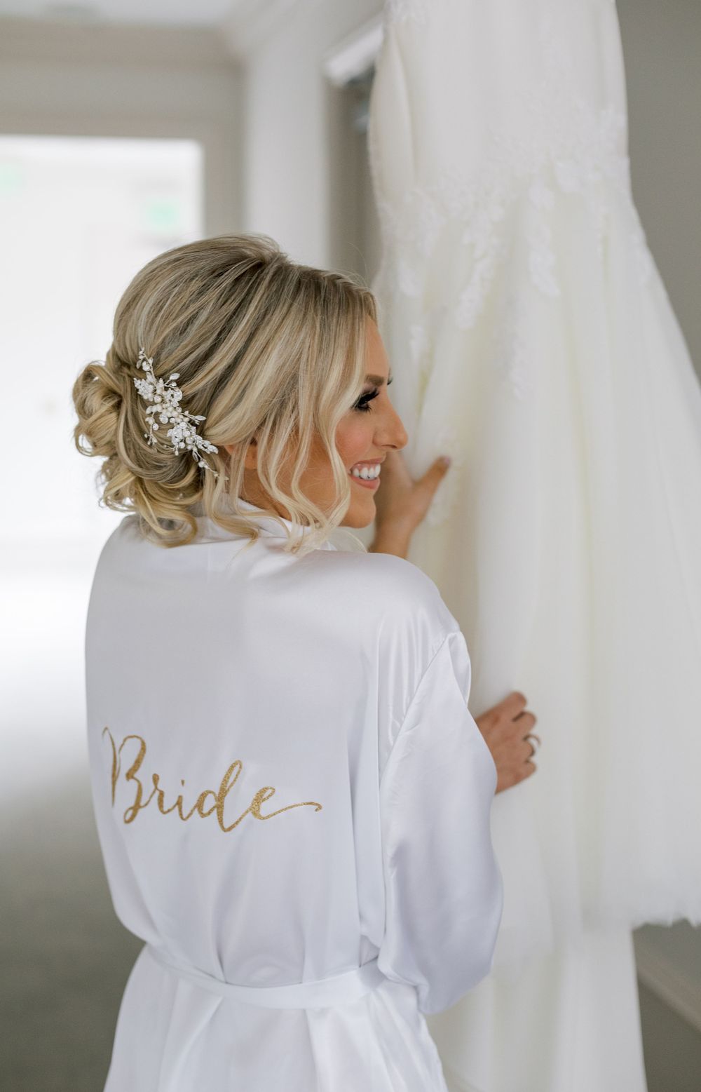 White silk robe with gold 'Bride' text shown with elegant wedding dress hanging in background.