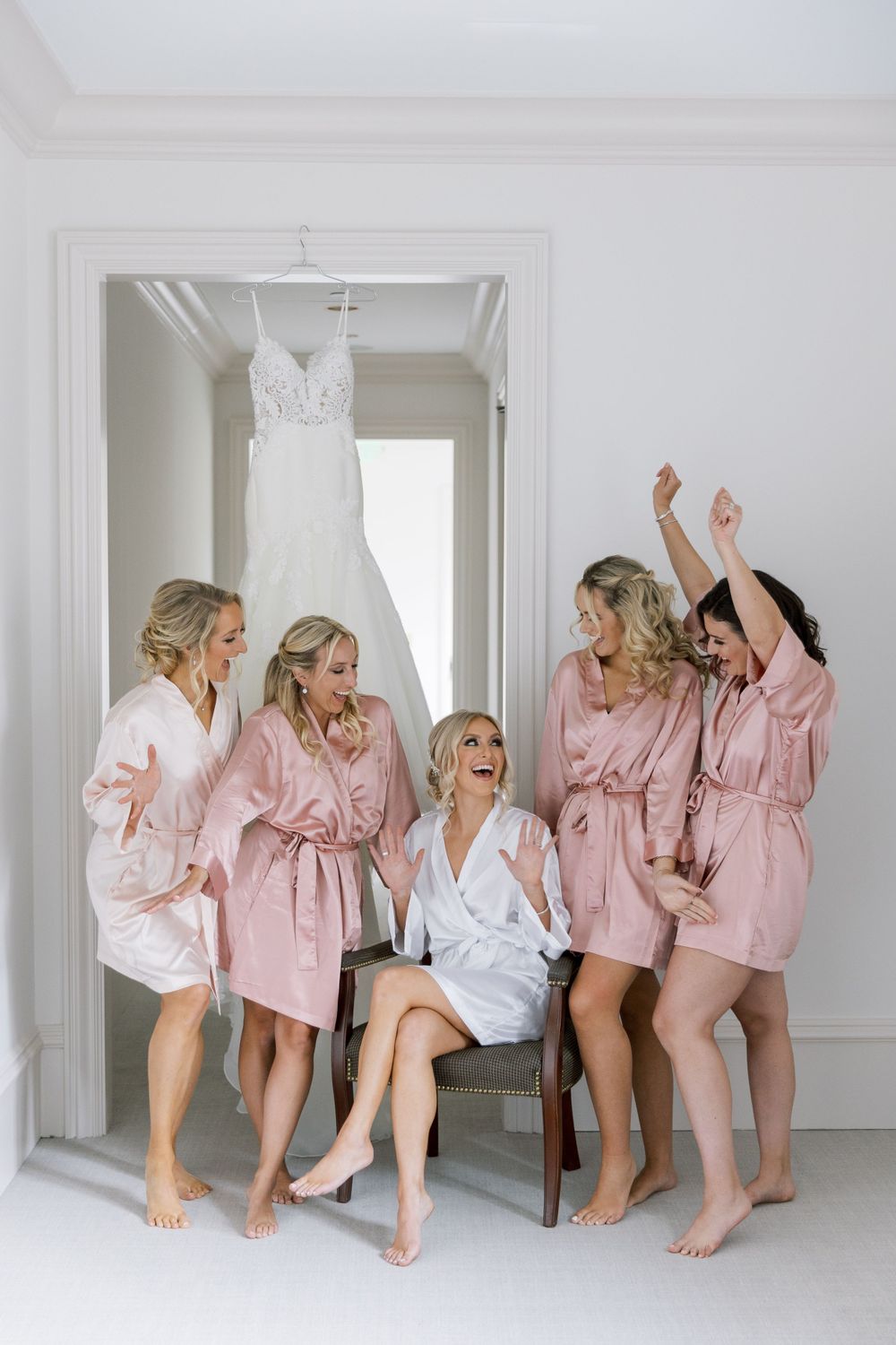 Group wearing pink and white silk robes celebrate with joy in front of hanging wedding dress.
