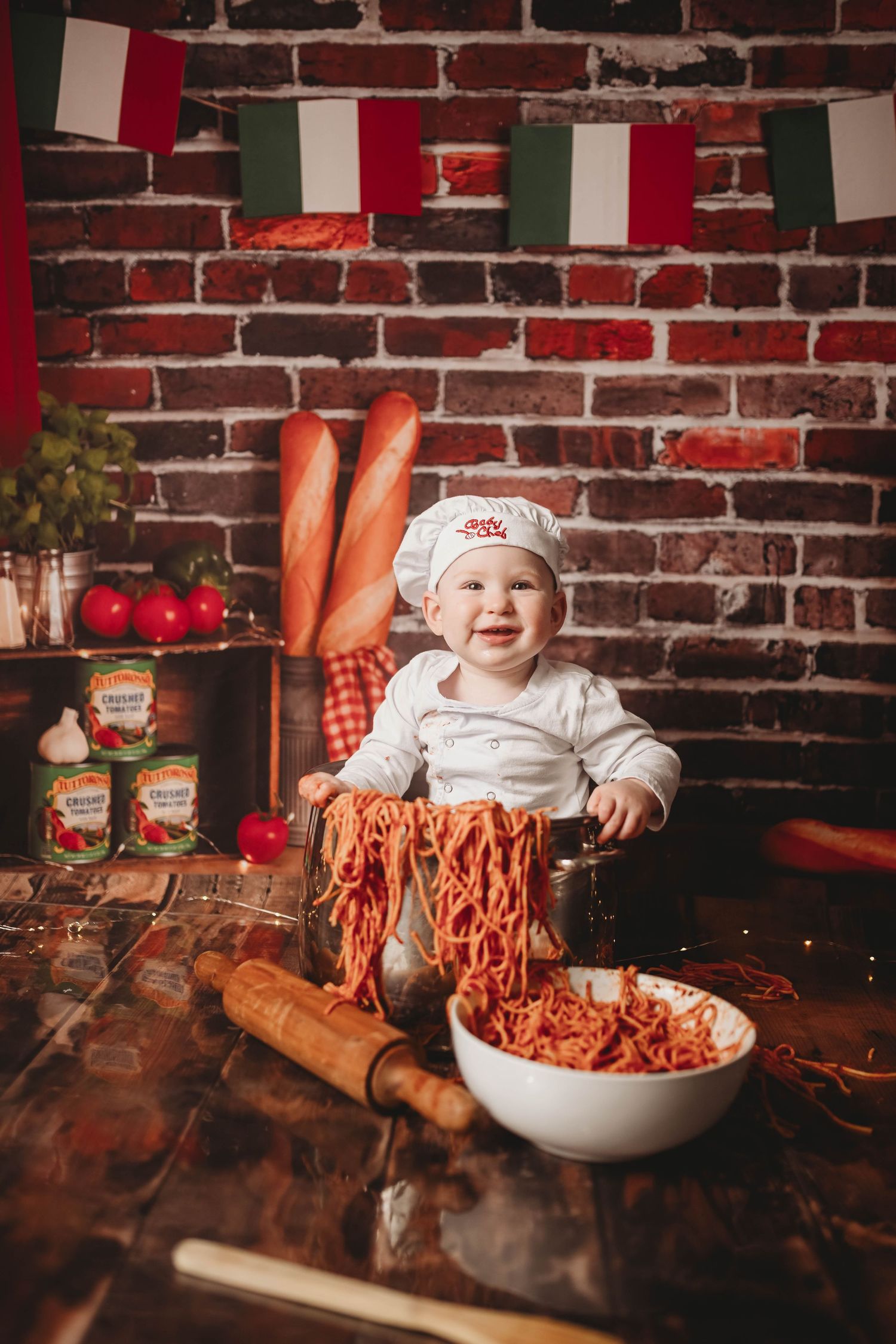 A young chef in a white hat plays with spaghetti and tomato sauce in a rustic Italian kitchen setting.