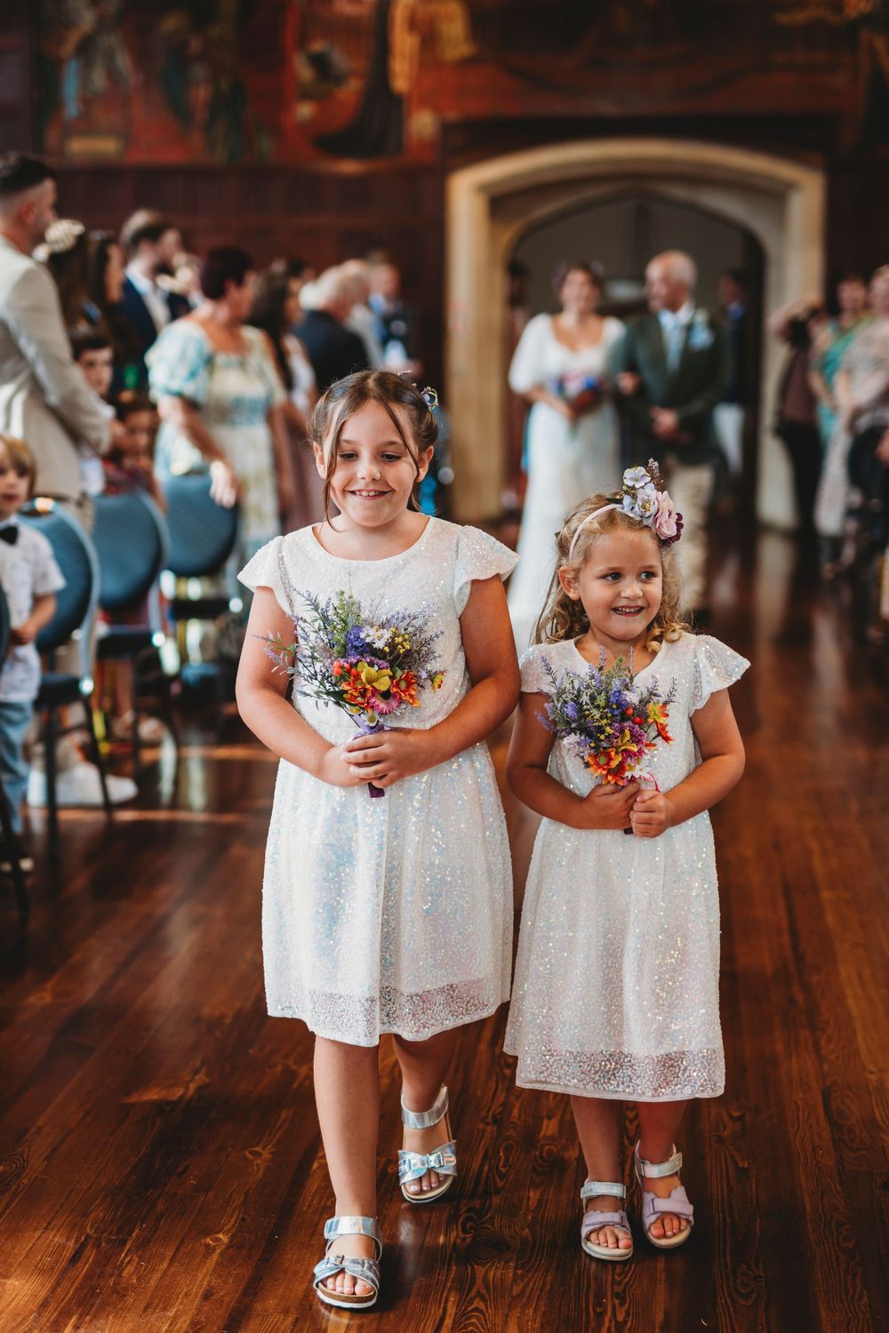 Young flower girls in white dresses hold colorful bouquets while walking on hardwood floors.