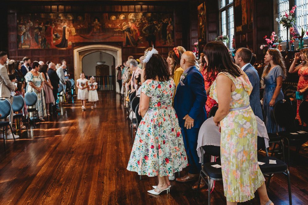 Wedding guests in floral dresses mingle in a beautifully decorated historic venue.