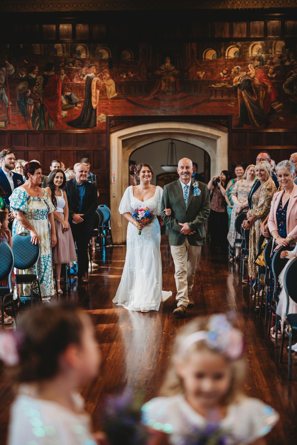 Wedding ceremony in progress inside an elegant venue with ornate wall murals and wooden floors.