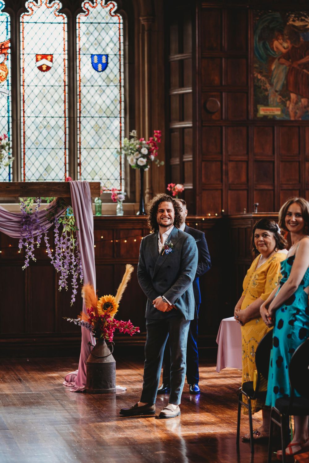 Wedding guests watch celebration in elegant wood-paneled room with stained glass windows.