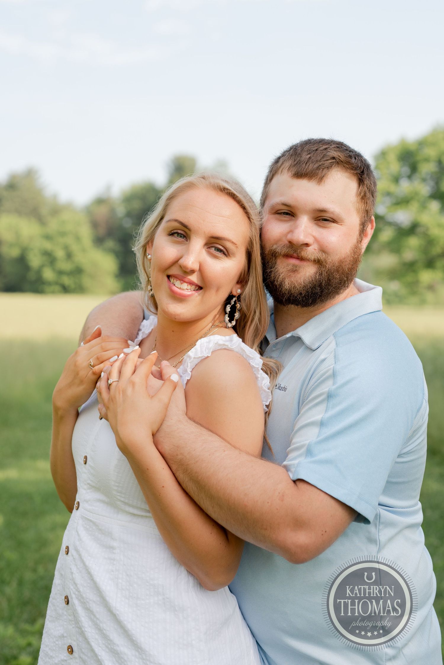 A joyful embrace captured in a sunny field with a natural backdrop of trees.