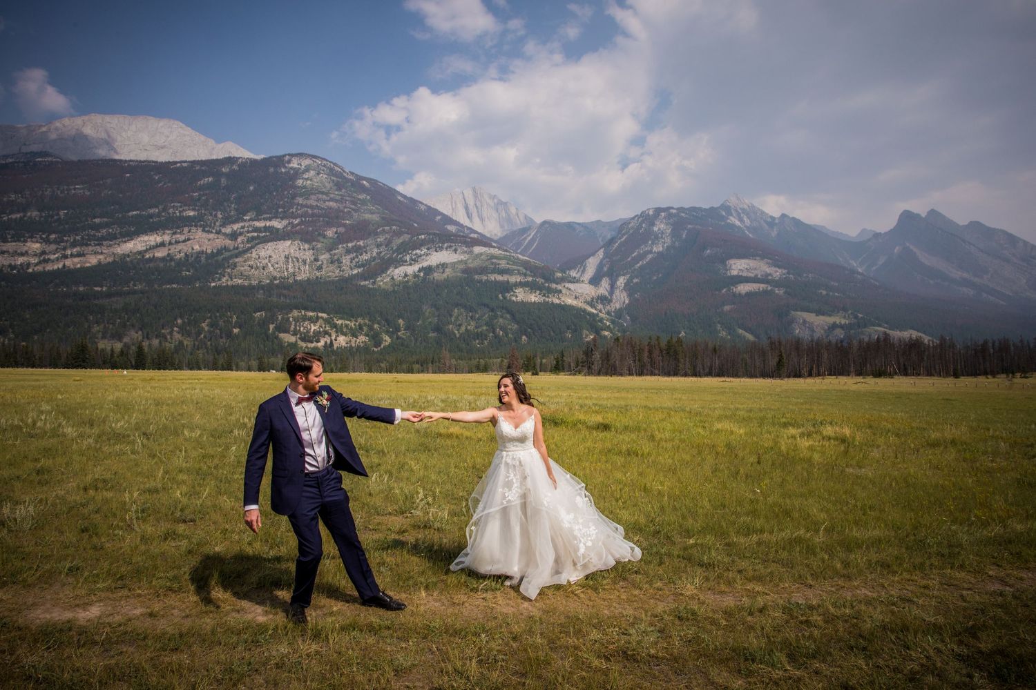 Couple in wedding attire poses playfully against dramatic mountain landscape backdrop in scenic meadow.