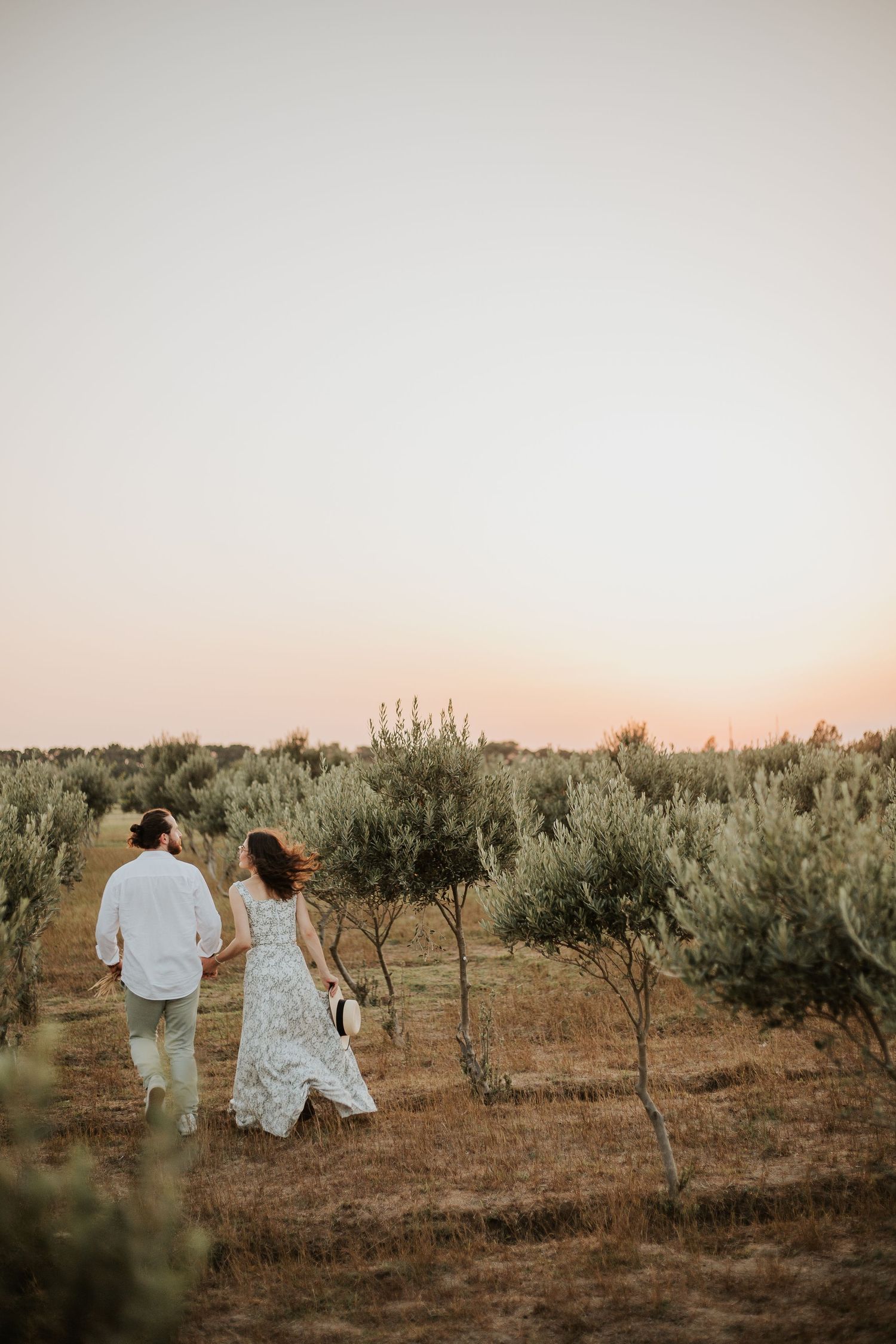 Couple walking through an olive grove at sunset in romantic wedding attire.