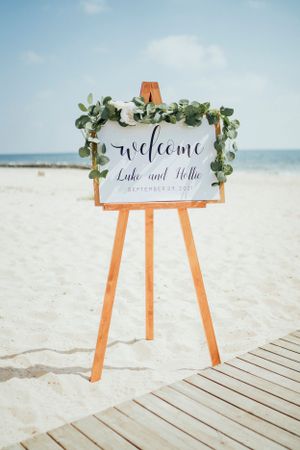 Wooden welcome sign with greenery decoration on an easel at a beach wedding.