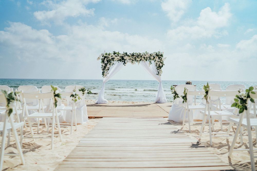 Beach wedding ceremony setup with white chairs and floral arch on a wooden boardwalk.