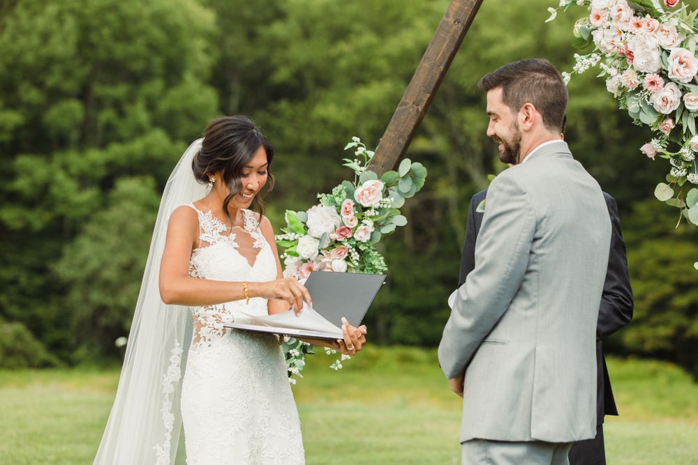 A wedding ceremony takes place outdoors with floral decorations and a couple exchanging vows beneath an arch.