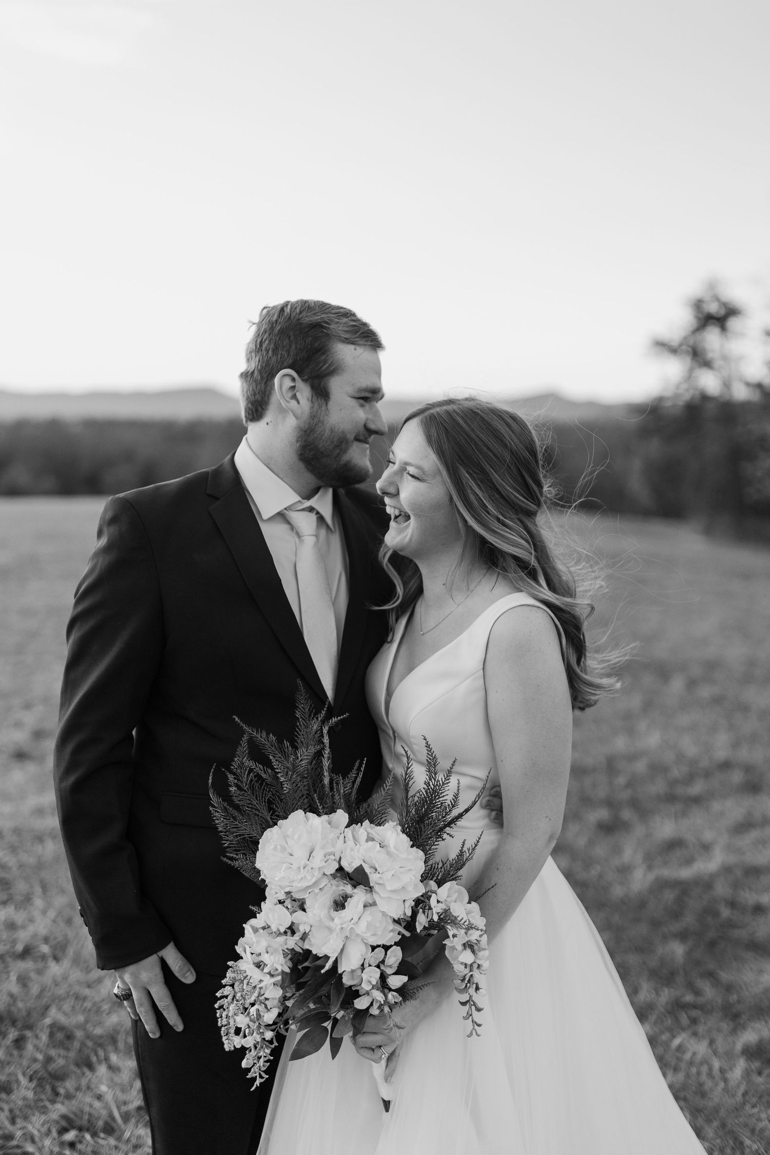 Black and white wedding portrait capturing joyful laughter between newlyweds with rustic bouquet.