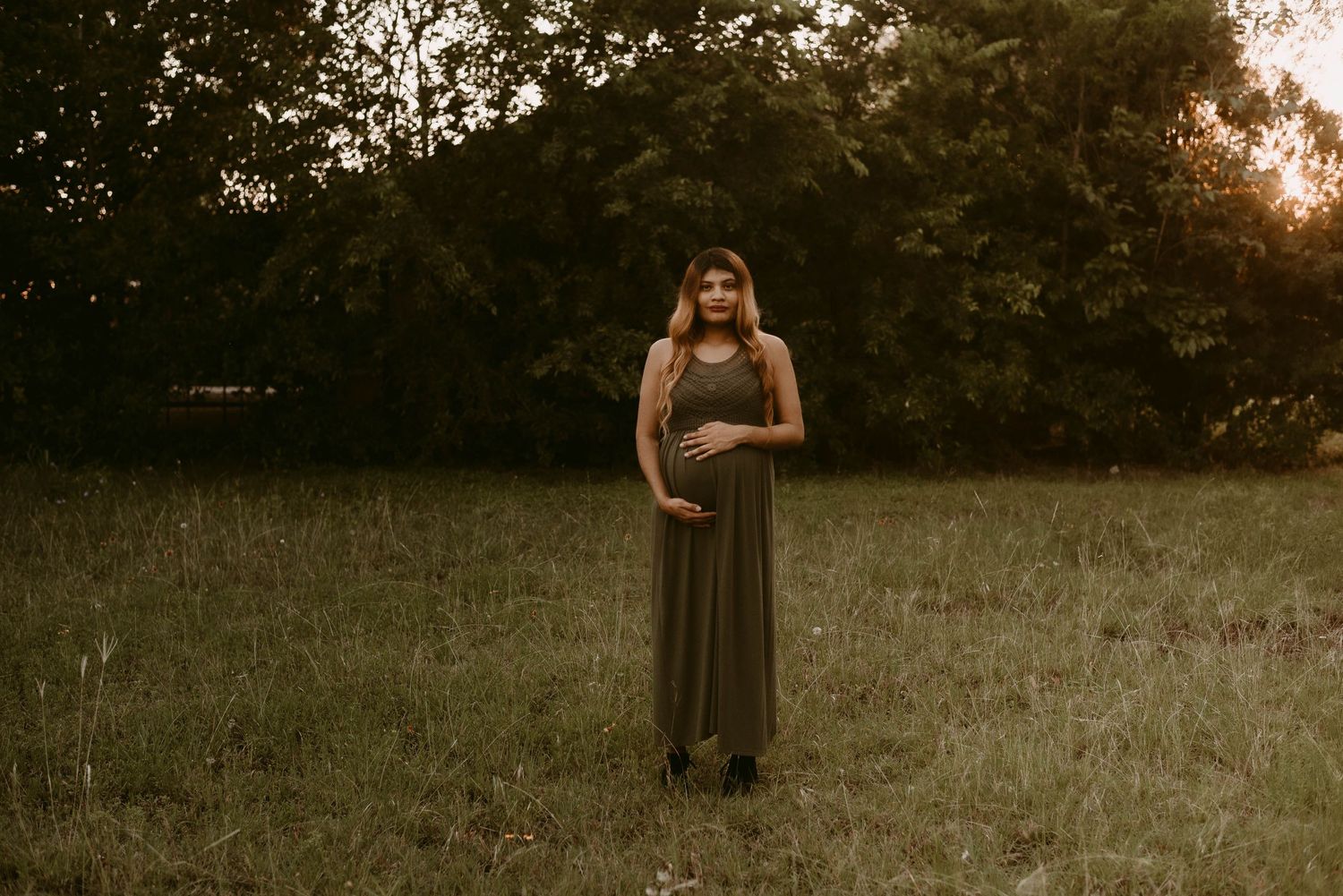 Expectant mother in long olive dress stands in sunlit meadow during golden hour portrait session.