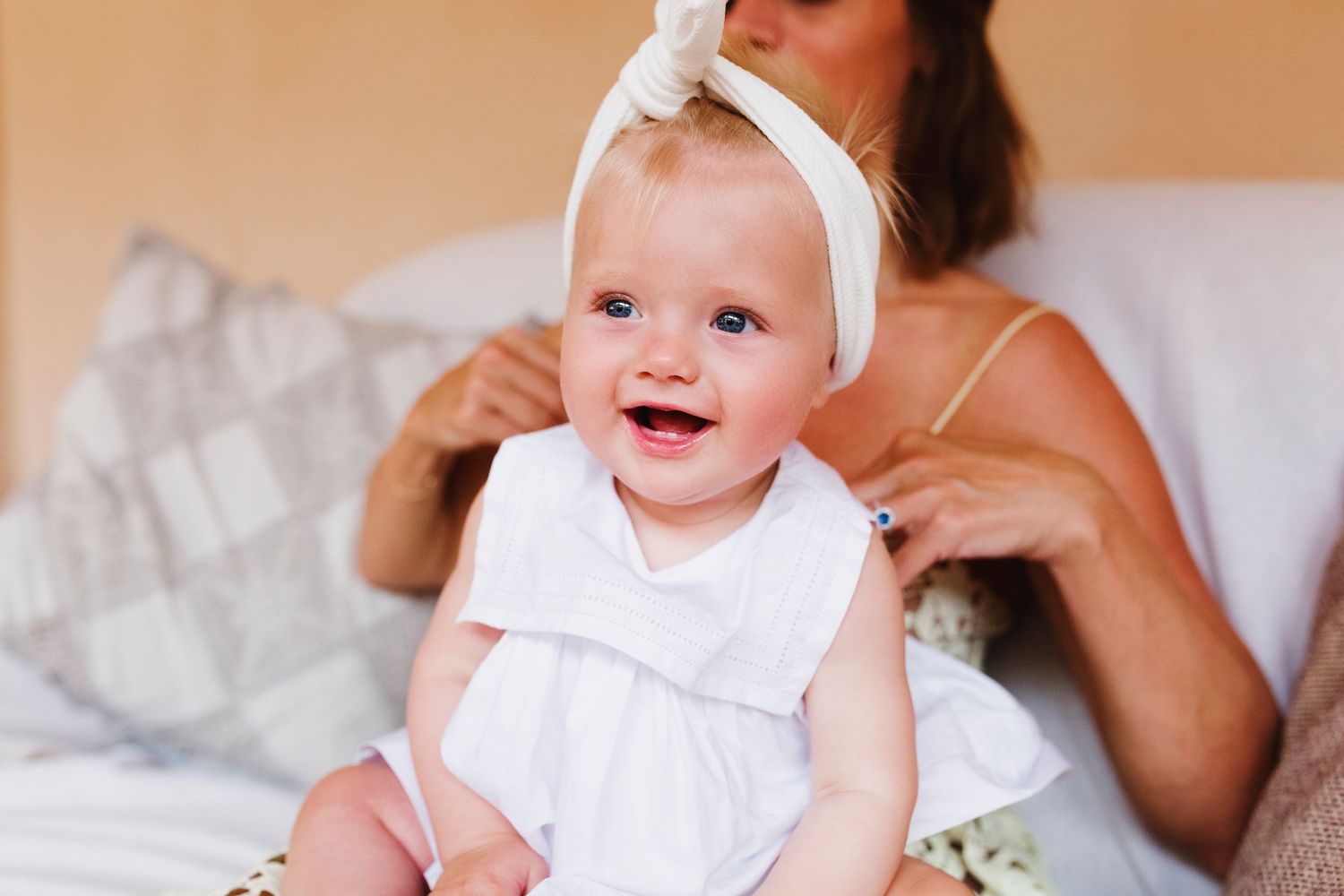 Baby wearing white dress and headband sitting on bed with joyful expression.