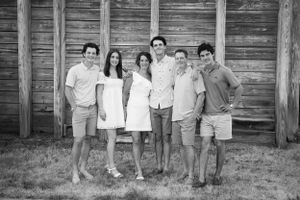 Group portrait of six young people standing together in casual summer attire against a rustic wooden wall.