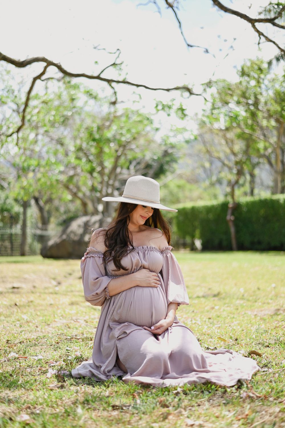 Maternity photoshoot in garden with flowing mauve dress and wide brimmed hat against natural backdrop.
