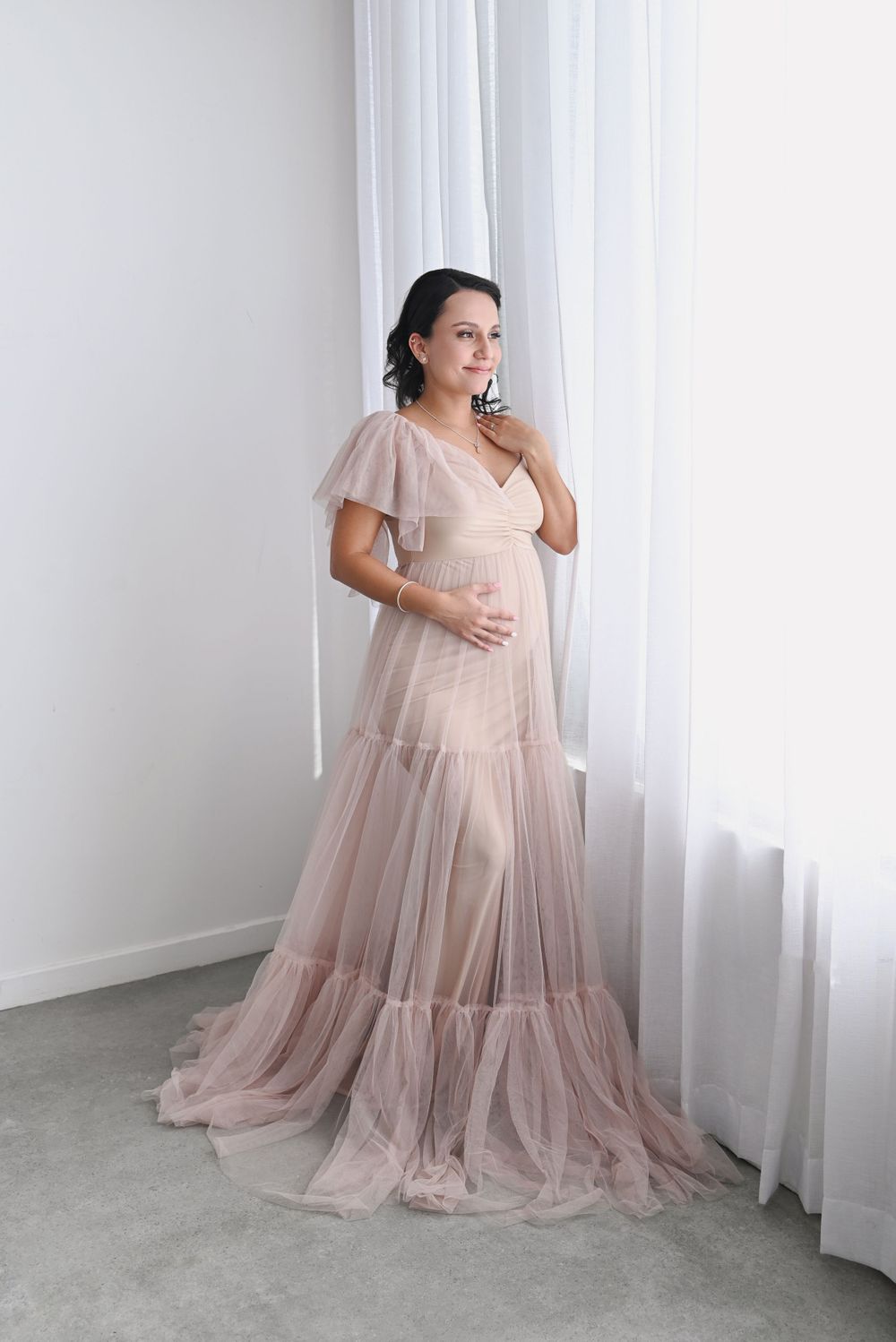 Ethereal maternity portrait in flowing blush pink gown against white curtained backdrop.
